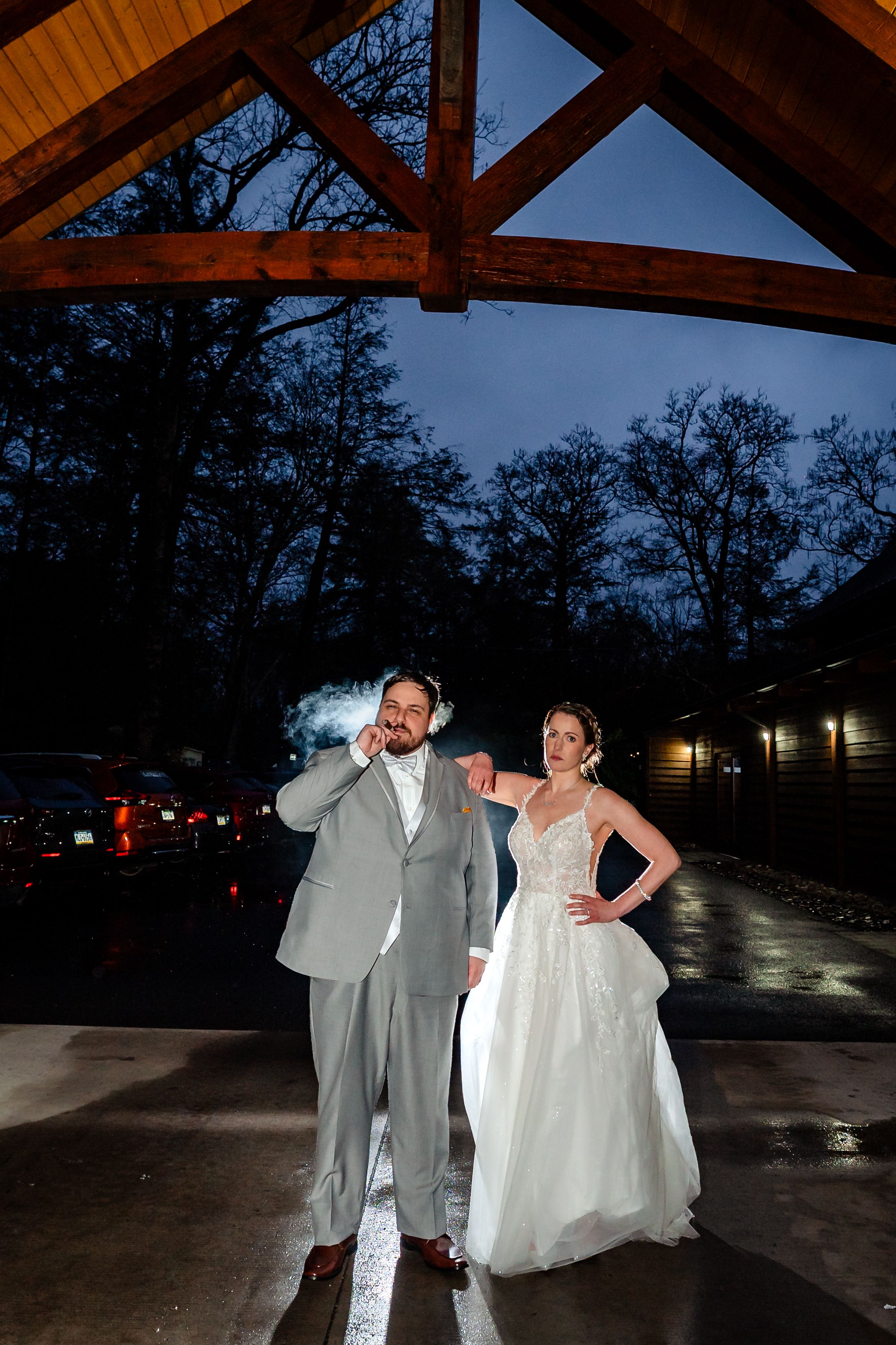 Bride and groom pose under a wooden pavilion at night, groom smoking a cigar in a gray suit beside his bride.