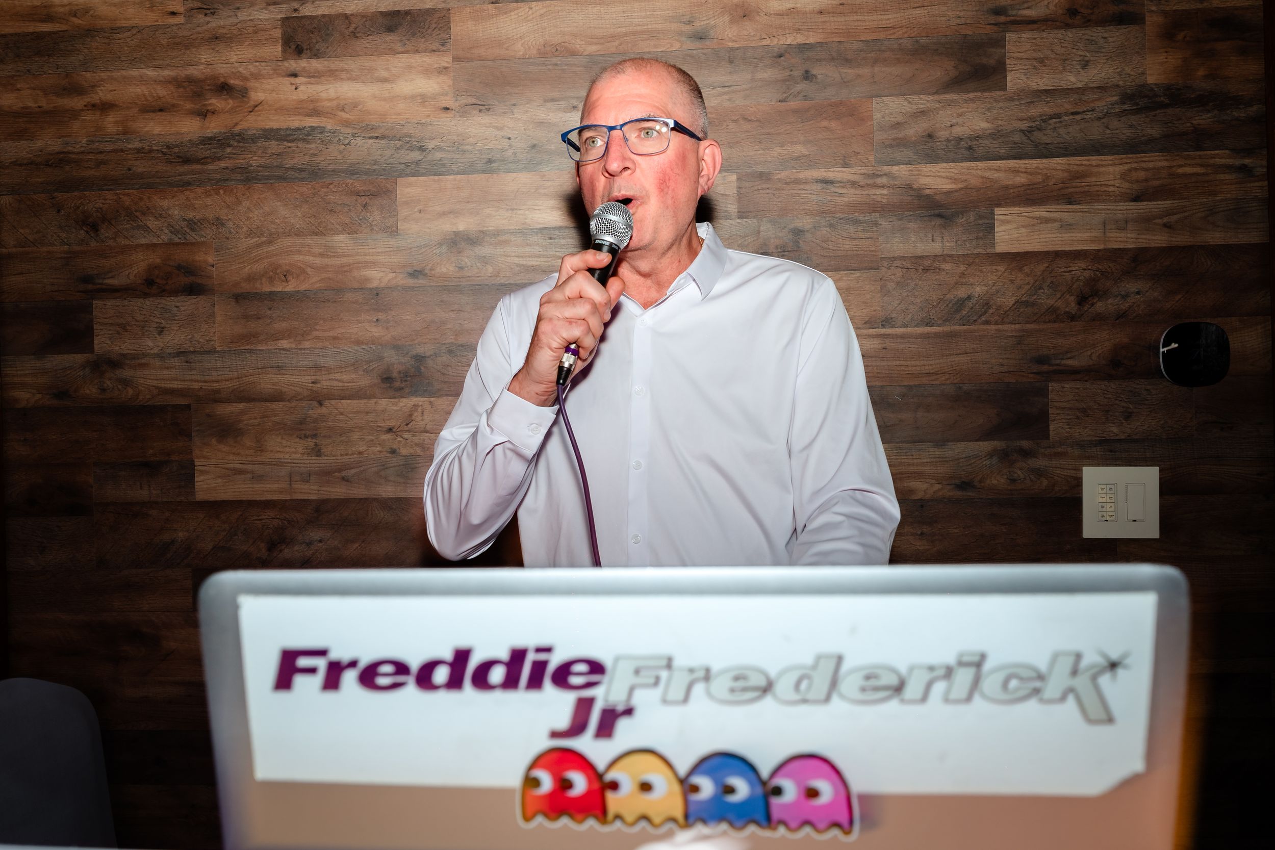 DJ Freddie Frederick Jr speaking into microphone at colorful Pac-Man themed DJ booth with wood panel backdrop.