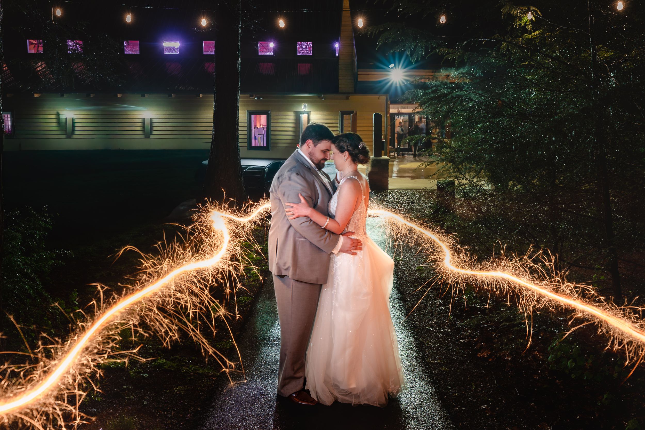 Newlyweds share a romantic kiss surrounded by glowing sparkler trails at night outside a warmly lit venue.