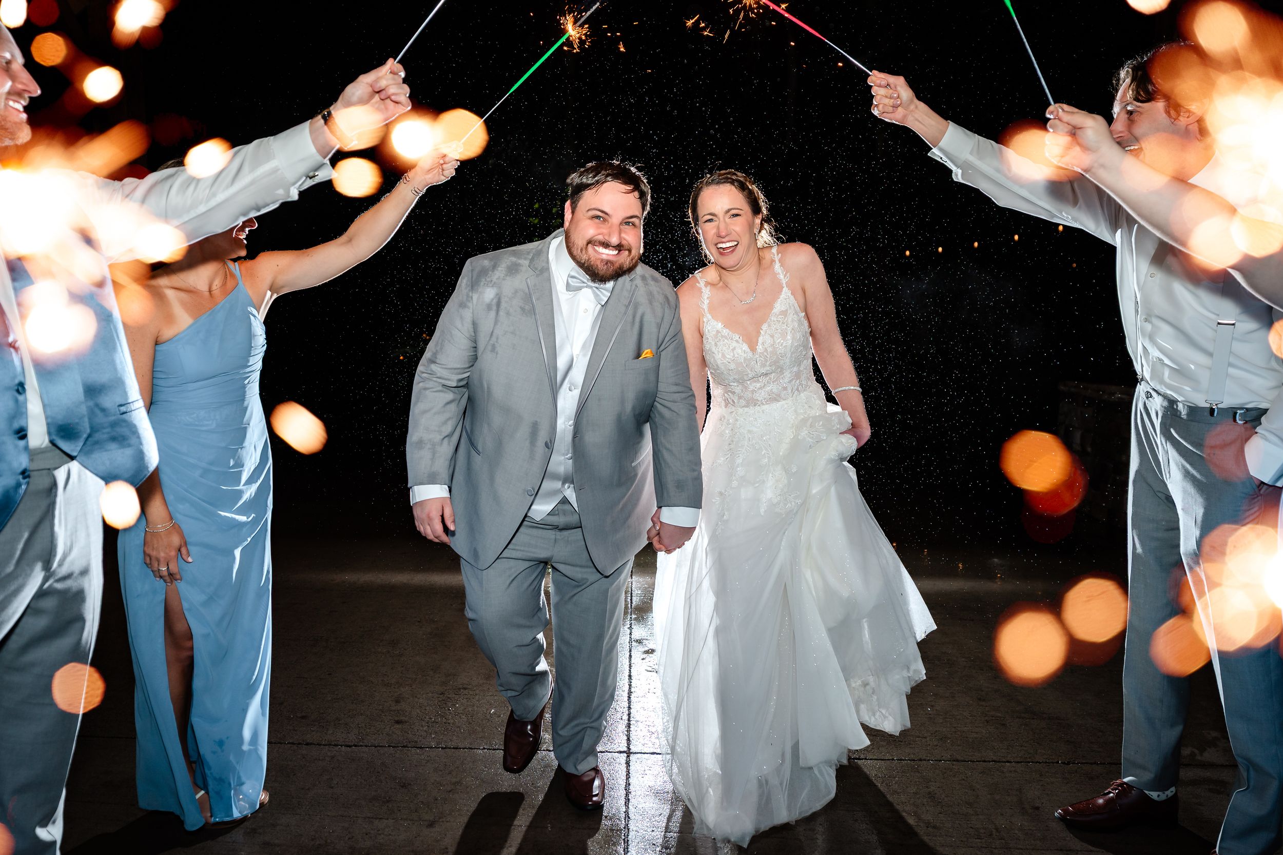 Bride and groom walk through a sparkler exit tunnel at night surrounded by wedding party members.