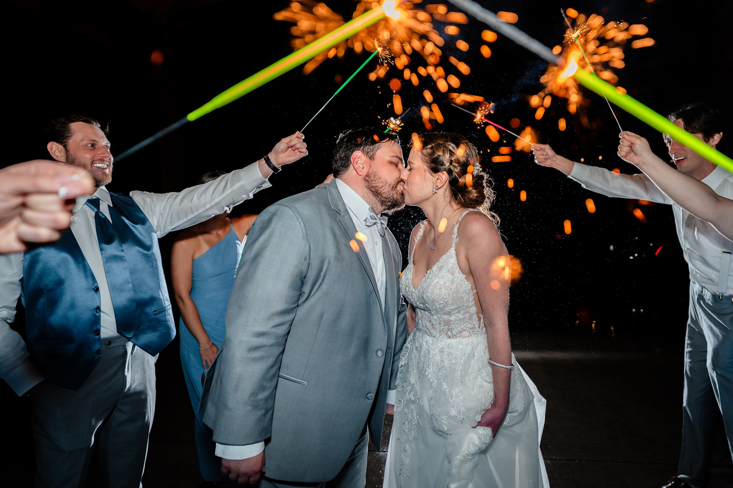 Newlyweds share a kiss surrounded by guests holding sparklers and glow sticks during a festive wedding send-off at night.