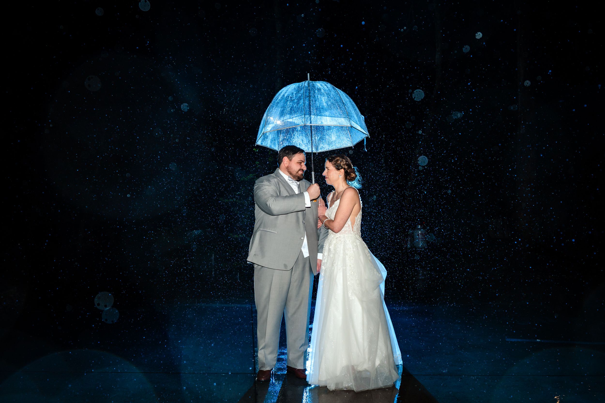 Bride and groom share a romantic moment under a glowing blue umbrella in the rain at night.