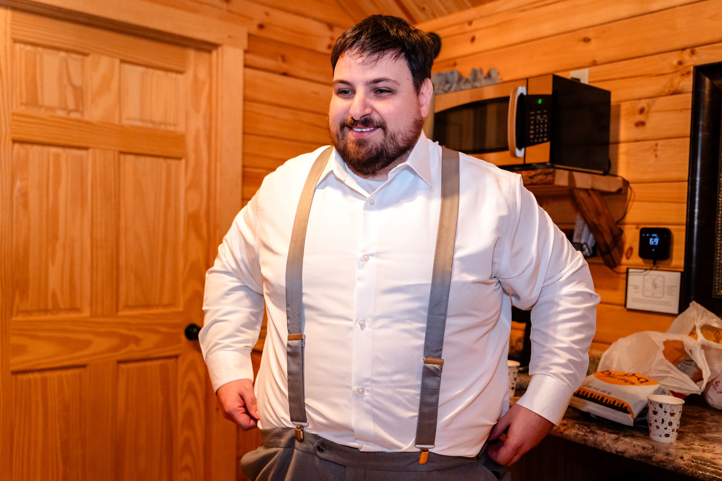 A smiling bearded groom in white shirt and gray suspenders poses in a rustic wooden cabin while getting ready.