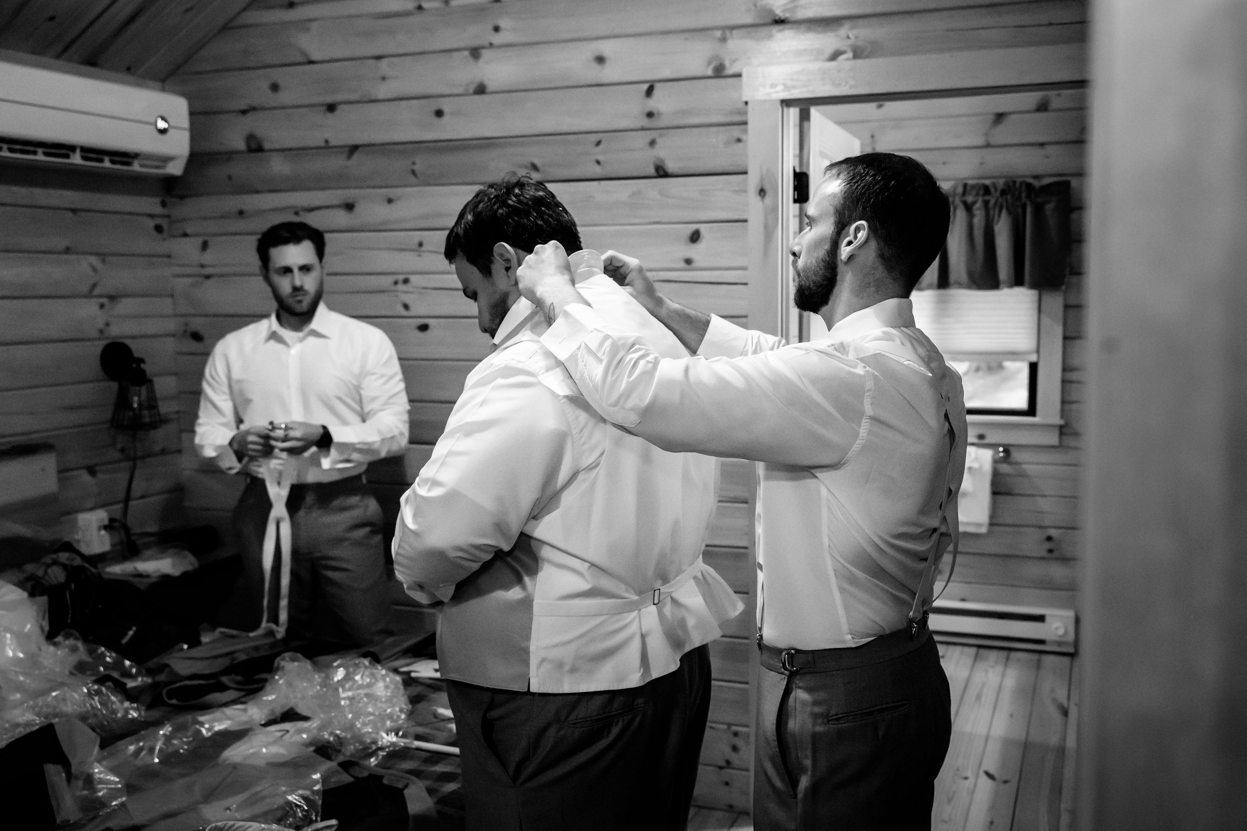 Groomsmen helping each other get dressed in a rustic wooden room before a wedding ceremony.