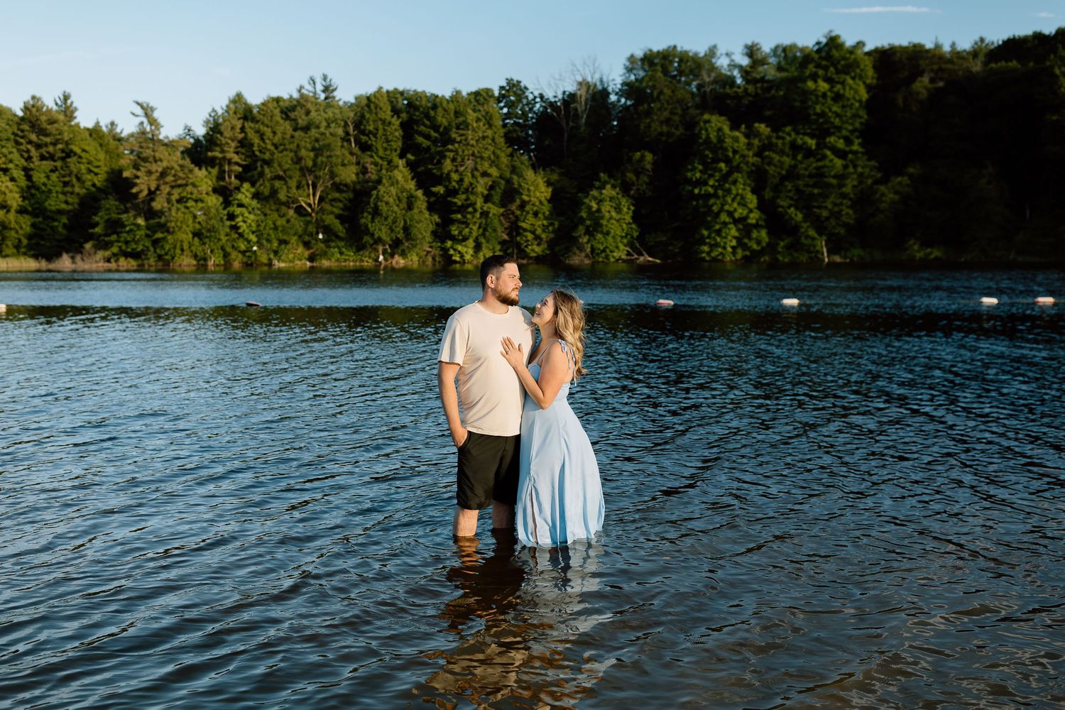 Couple standing in a lake with lush green trees, wearing casual summer attire, sharing a romantic moment outdoors.