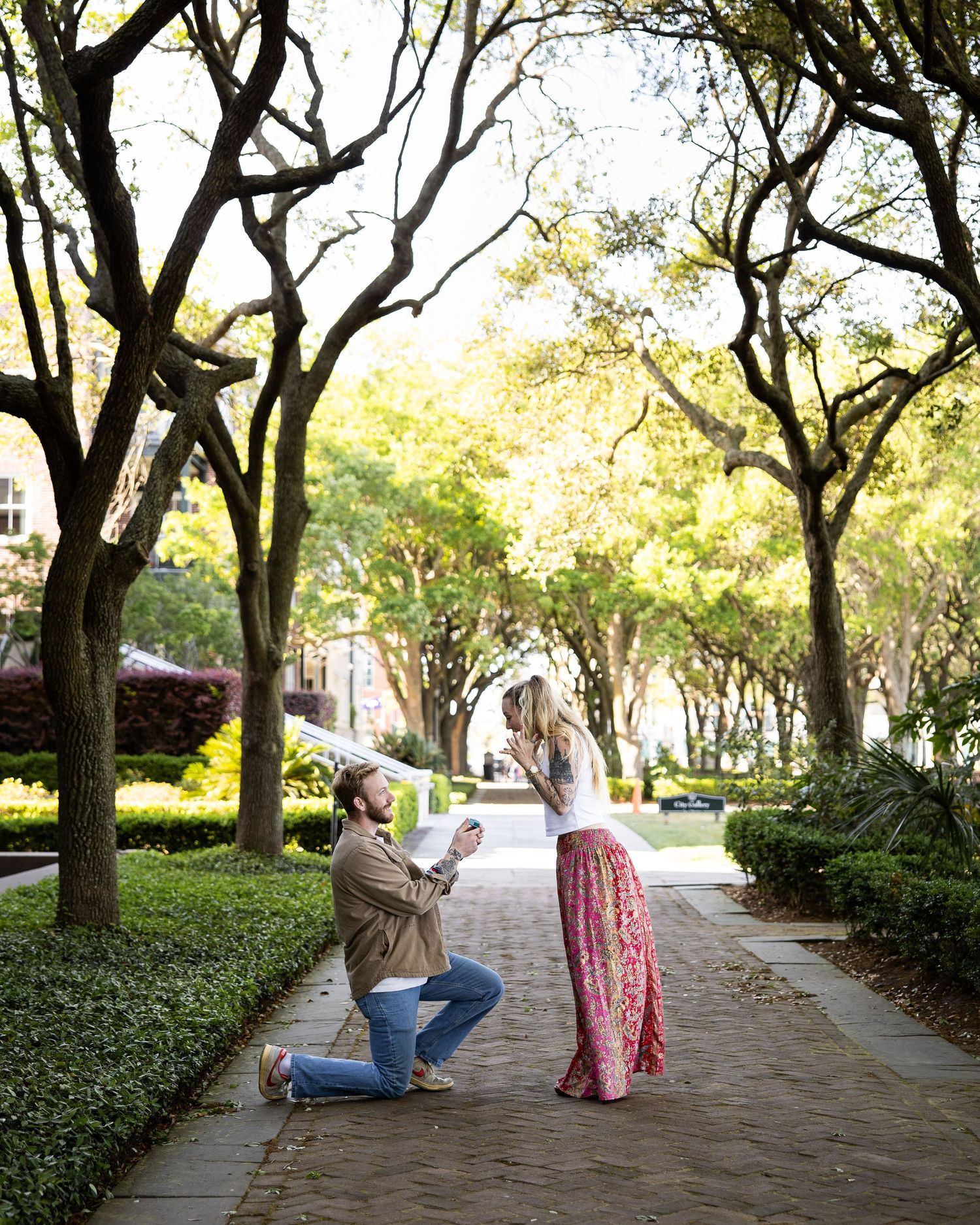 Man proposing to woman on one knee in tree-lined park walkway with brick path and green hedges.