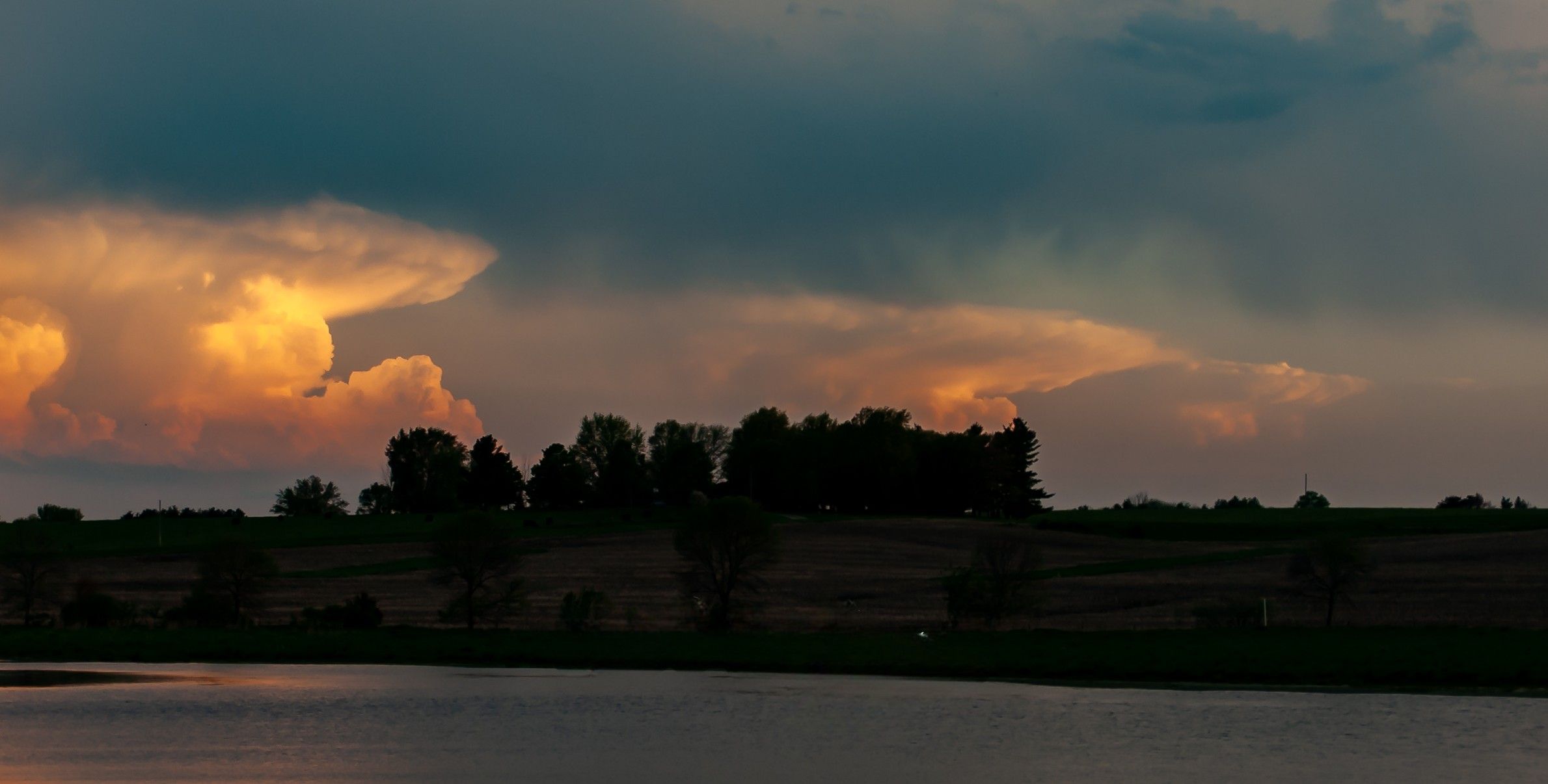 Thunderstorm clouds lit golden at sunset over lake and treeline Northern Missouri