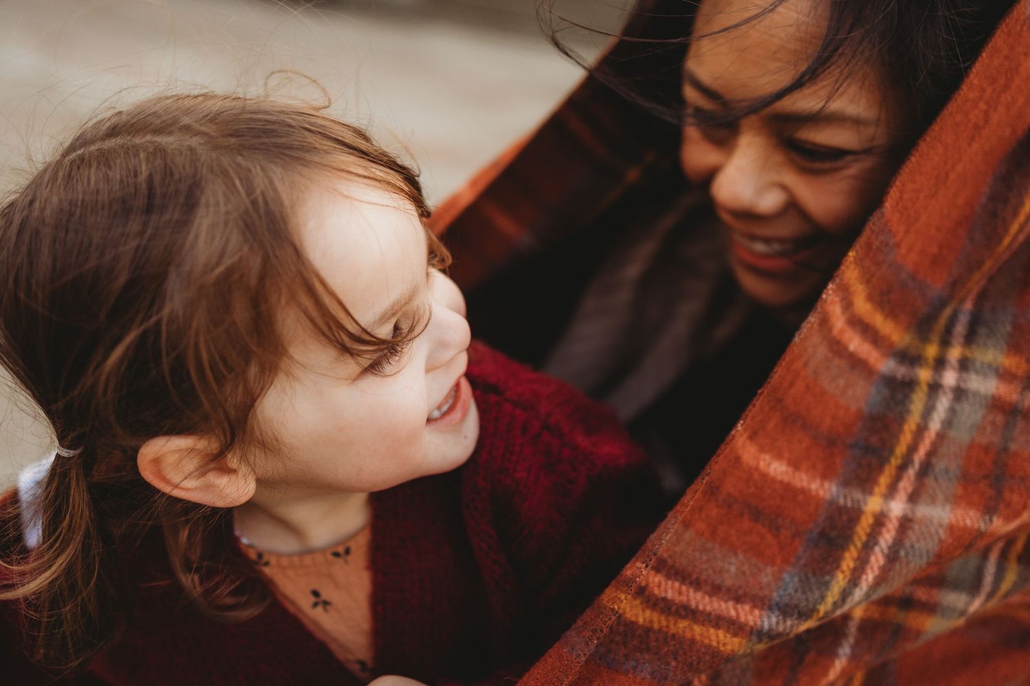 A young girl and woman share a warm moment wrapped in a cozy plaid blanket, smiling at each other.