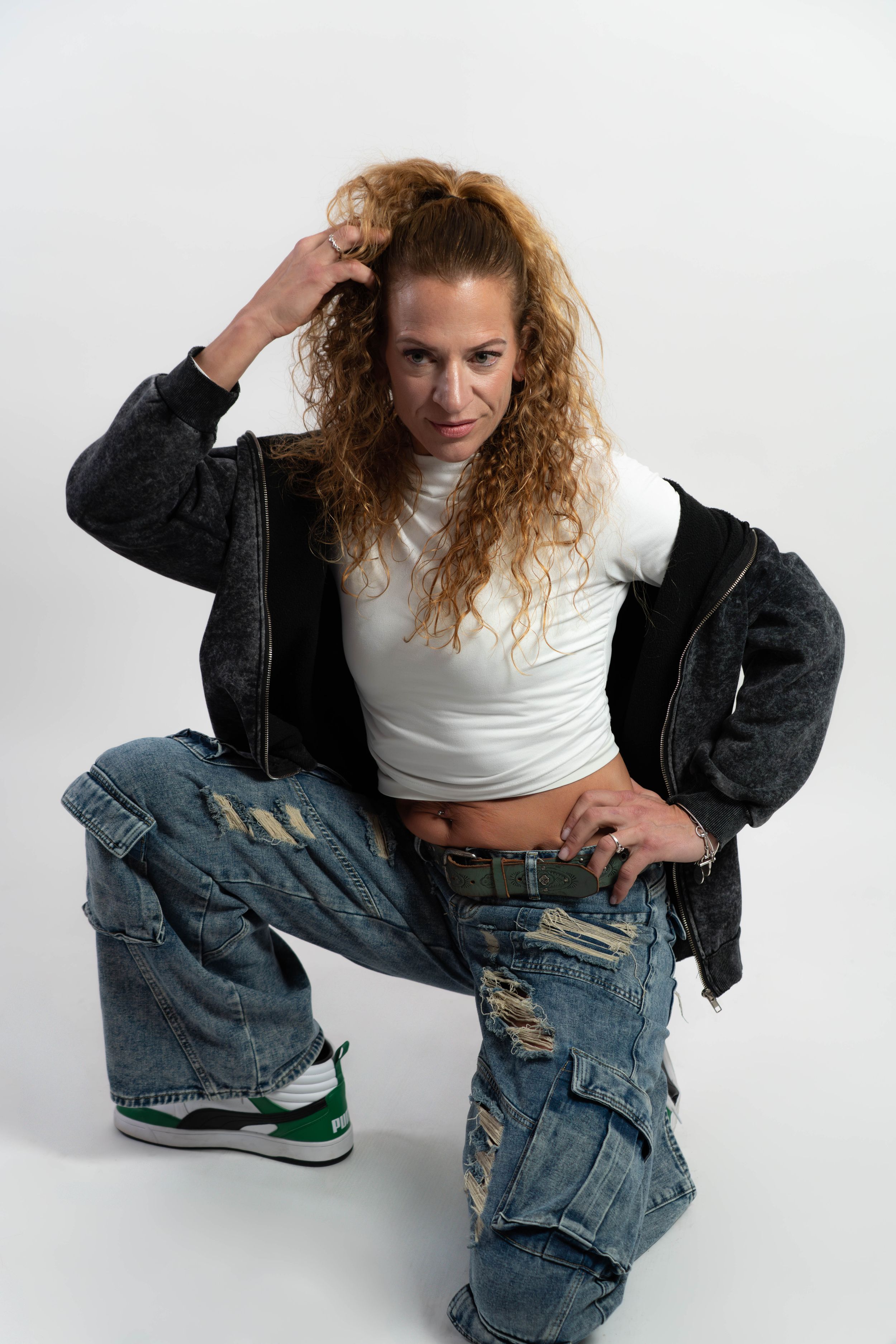Woman in ripped baggy jeans, white crop top, and dark jacket posing confidently on white background.