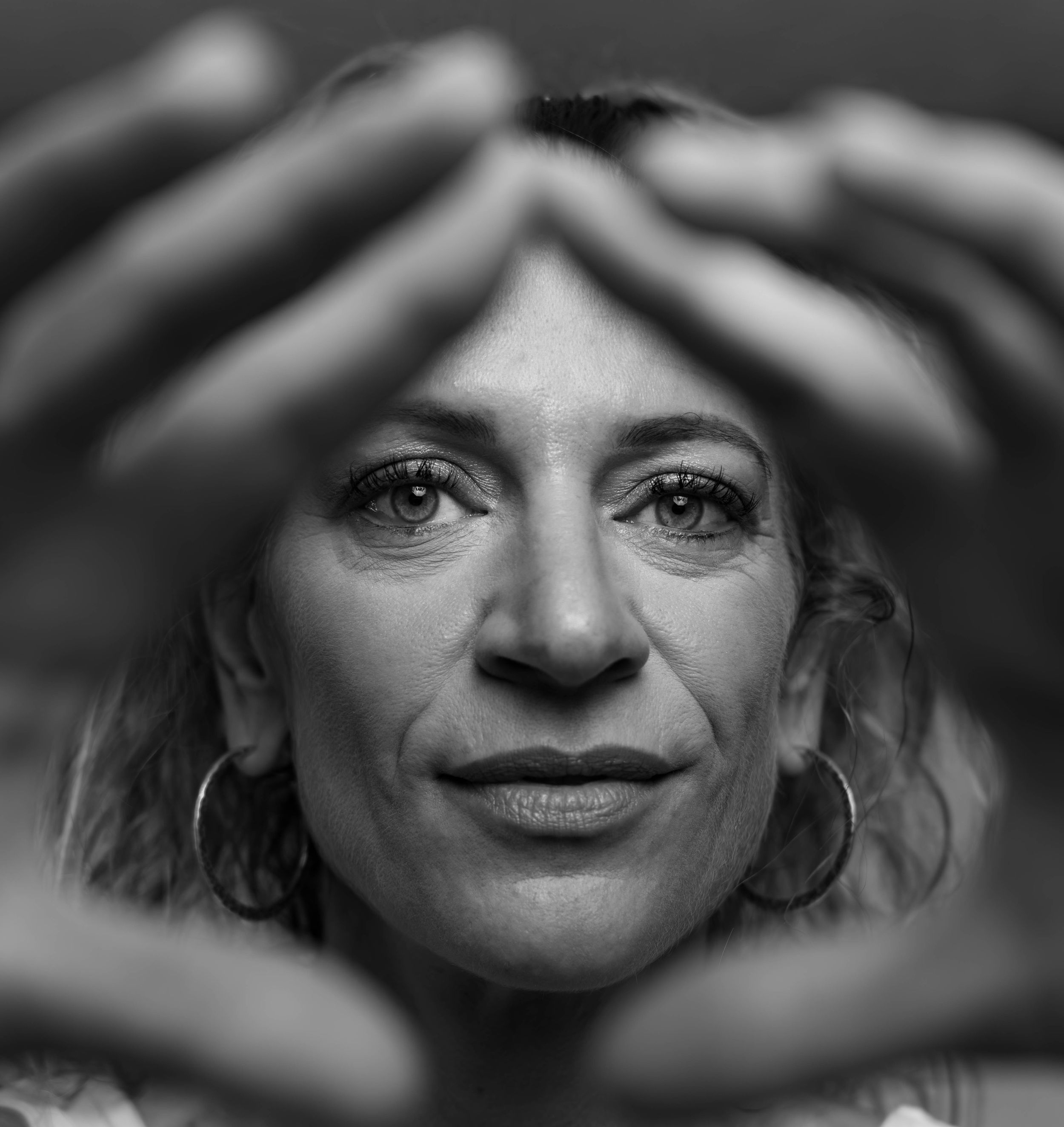 Black and white portrait of a woman framing her face with both hands, wearing hoop earrings, looking directly at camera.