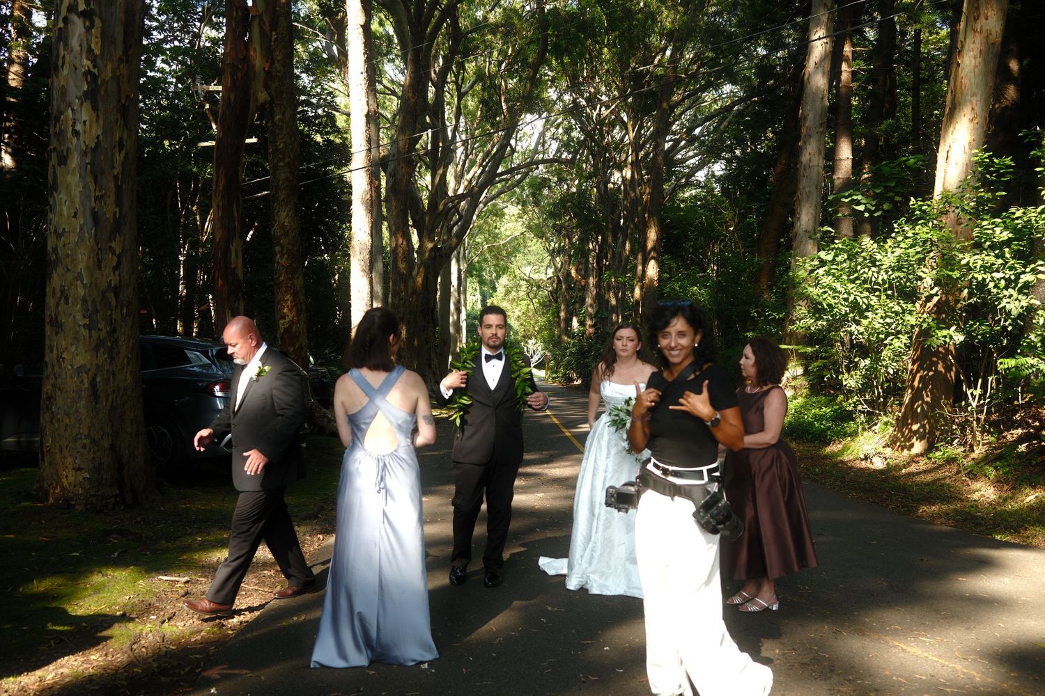 Wedding party walking along a tree-lined path, with bridesmaids in blue and burgundy dresses outdoors.