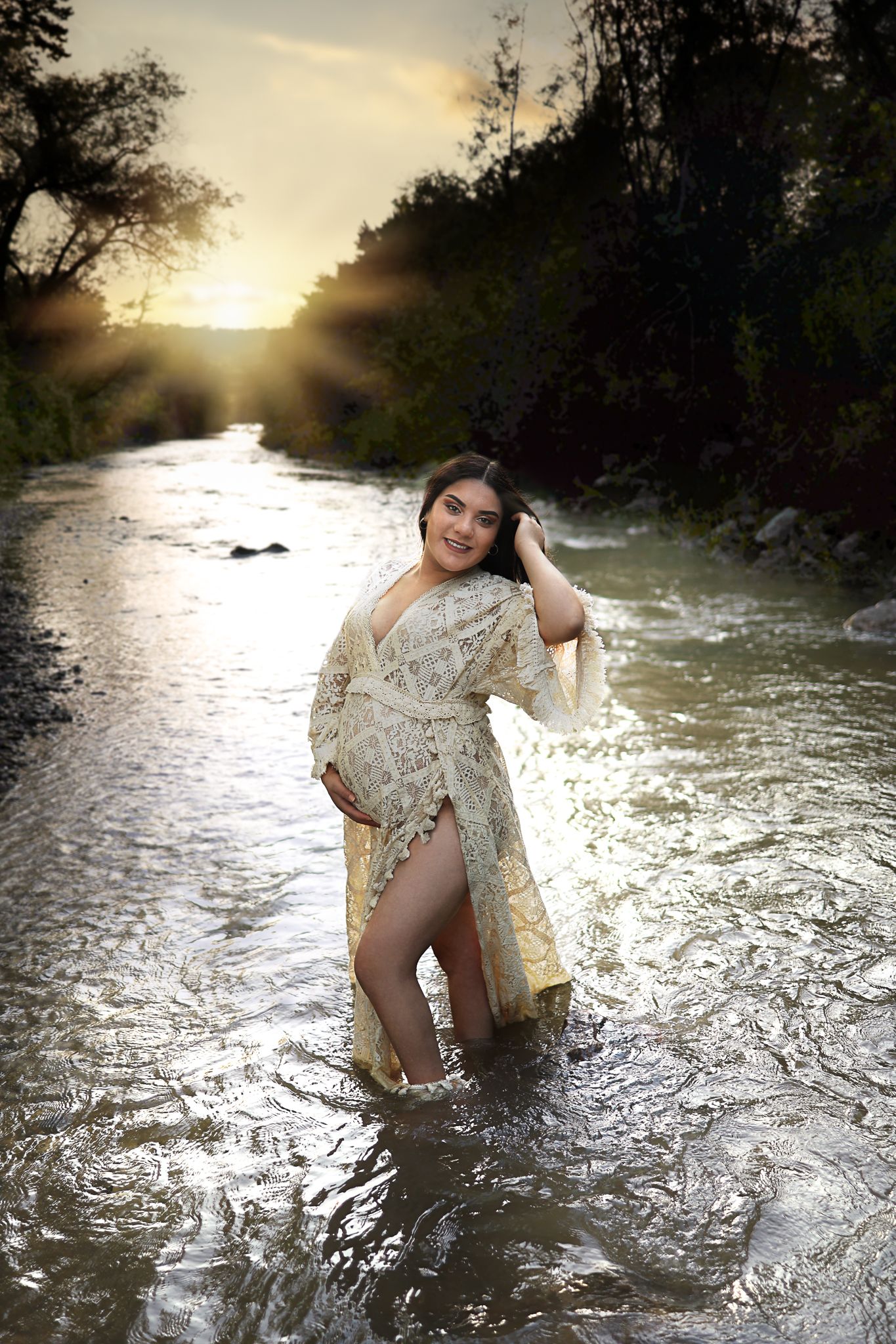 Pregnant woman in lace robe standing in a flowing river at golden sunset surrounded by lush greenery.