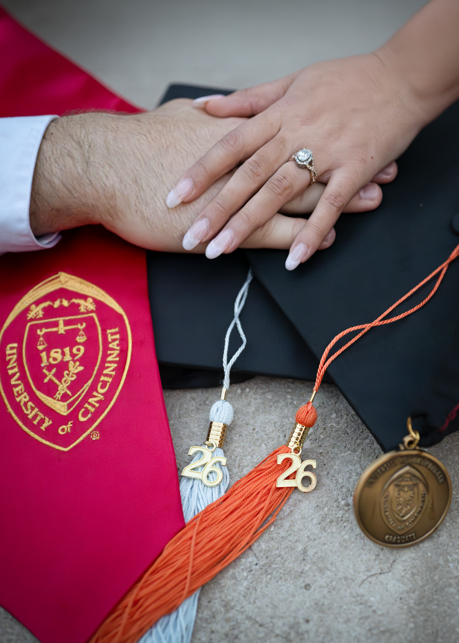 Two hands with an engagement ring rest on a University of Cincinnati graduation cap with Class of 2026 tassels and medal.
