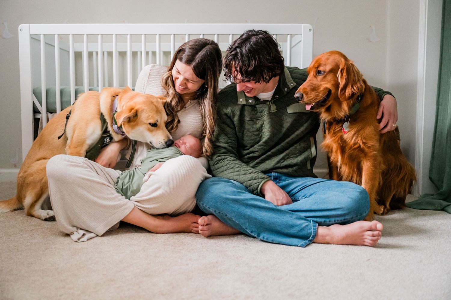 Connecticut family with newborn baby sitting on floor with two dogs, a golden retriever and a red setter, near a white crib.