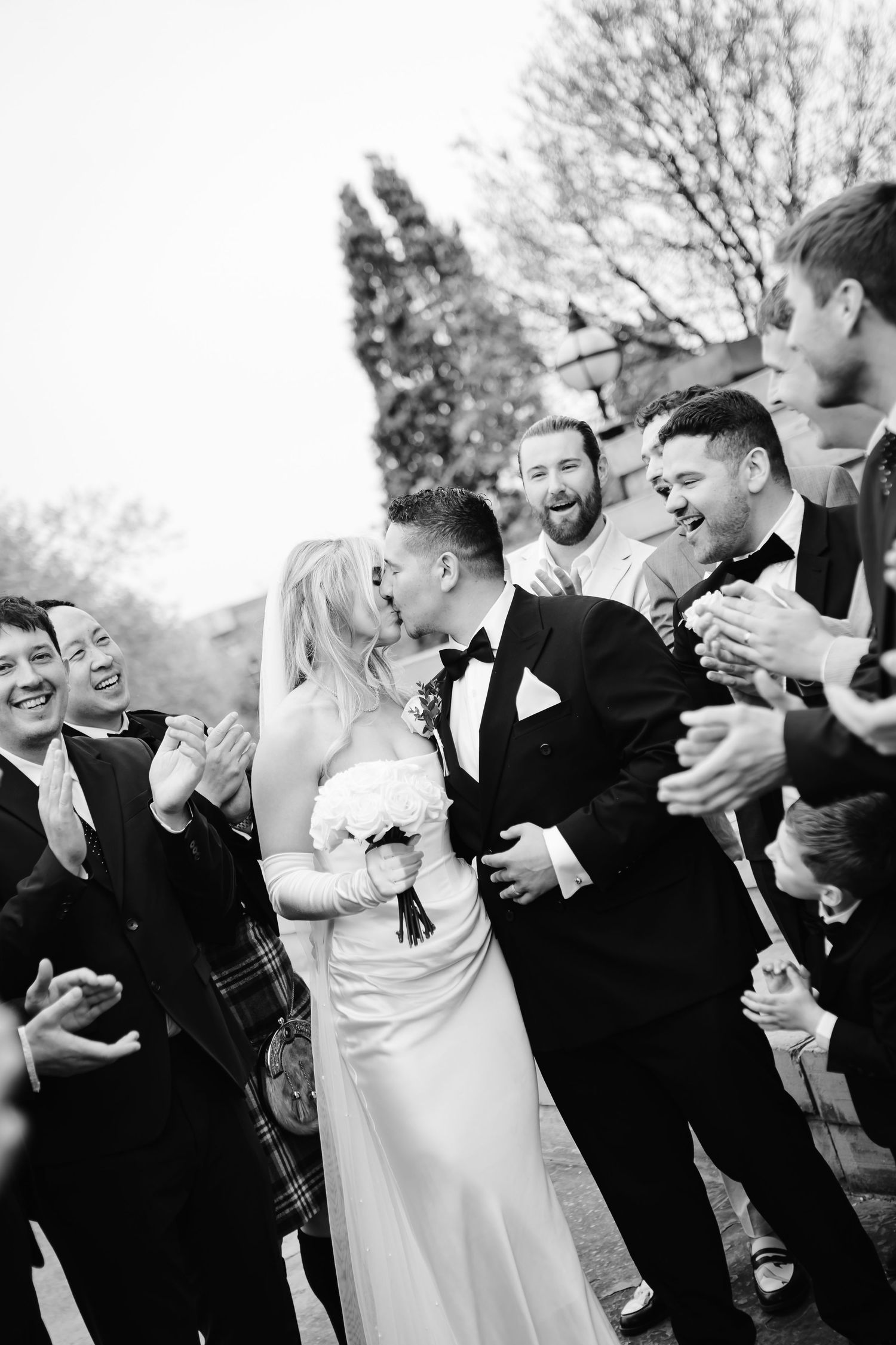 Bride in white gown and groom in tuxedo walk through cheering wedding guests outdoors.