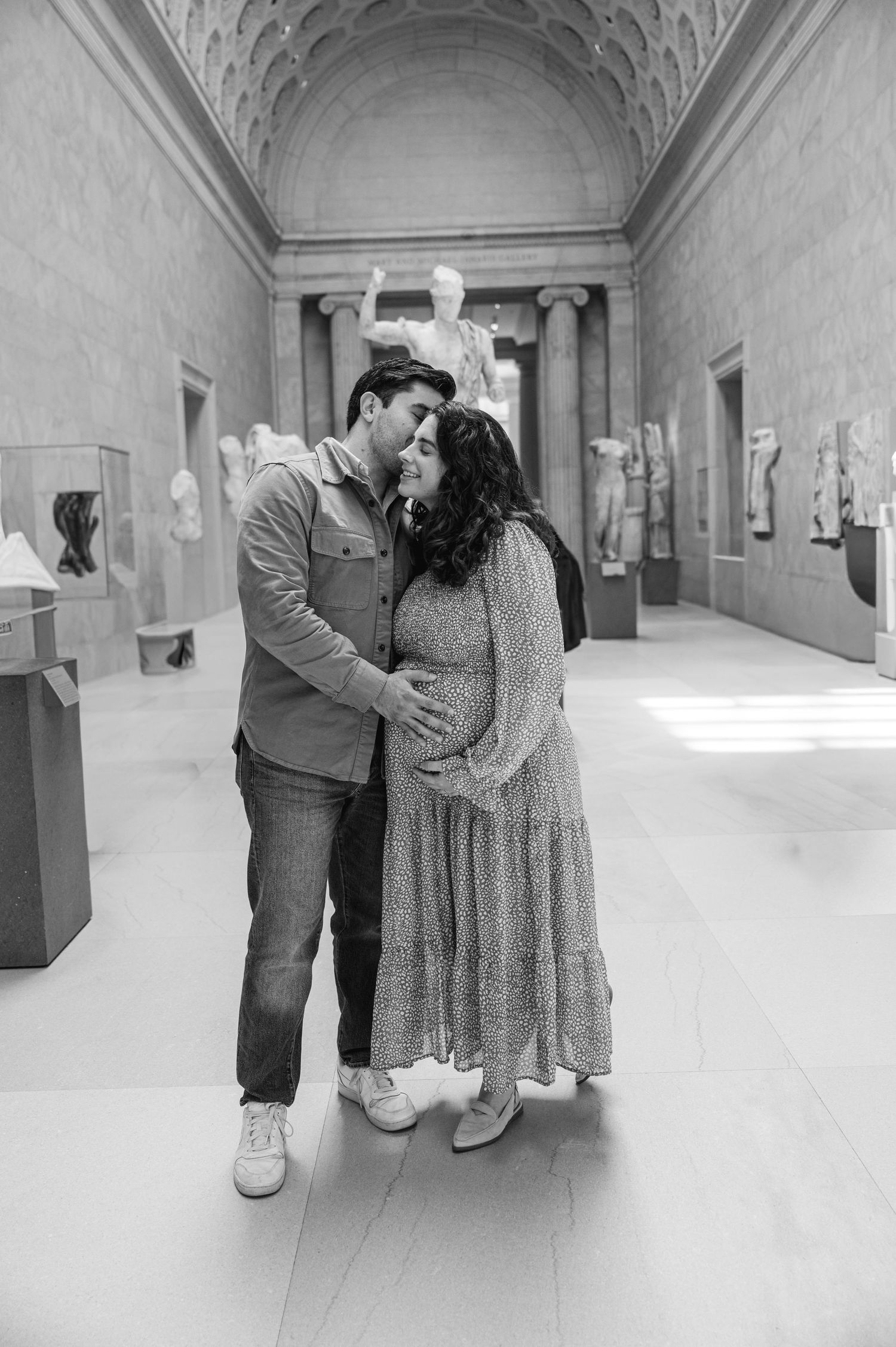 Black and white maternity photo of a couple embracing in the Greek gallery of the Met Museum in NYC surrounded by classical sculptures.