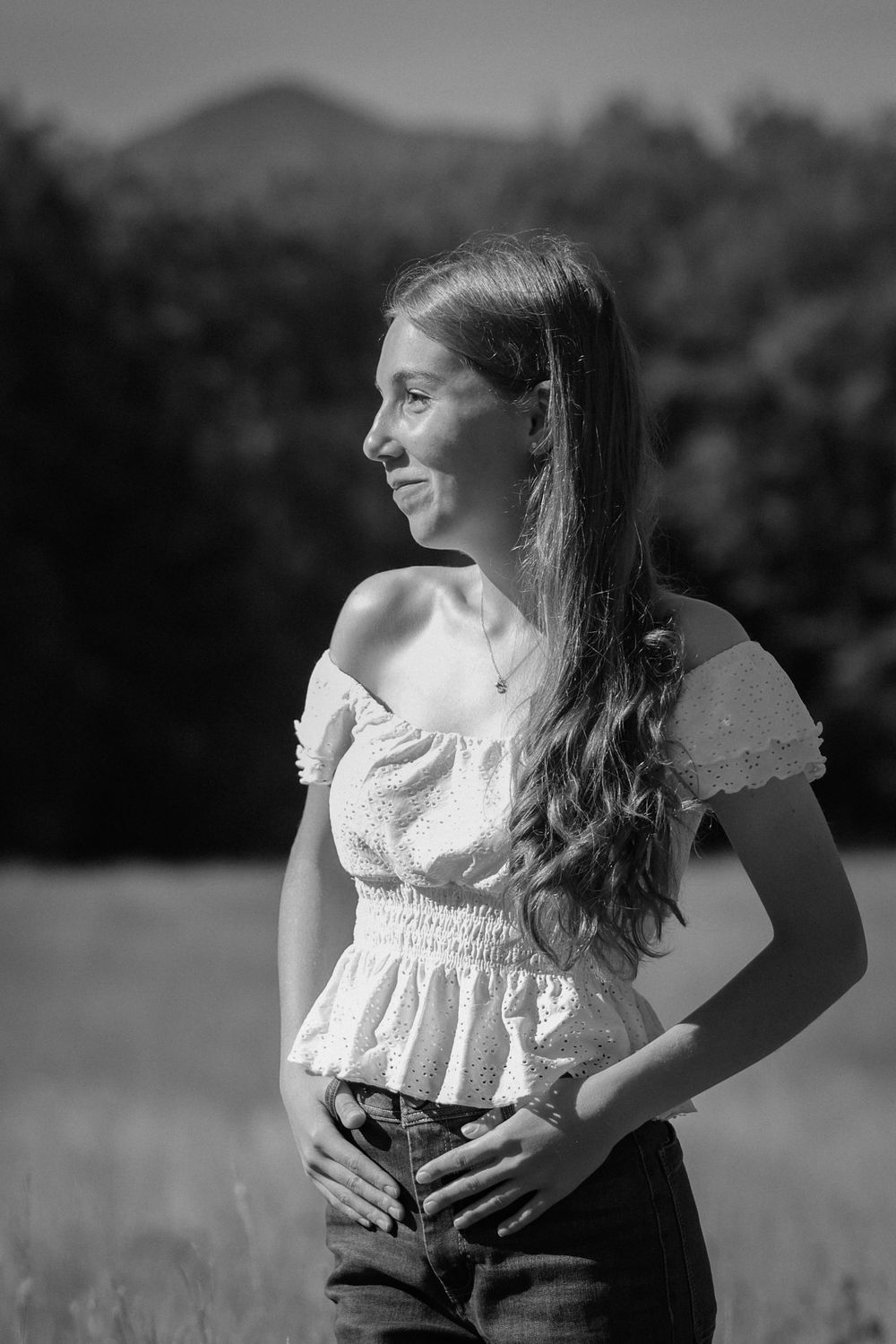 Black and white portrait of a smiling young woman in an off-shoulder top standing in a field.