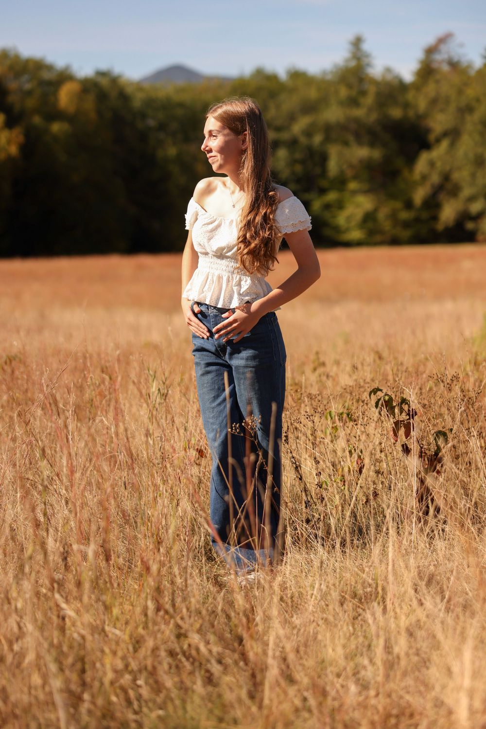 Young woman in a white ruffled top and jeans standing in a golden field with mountains behind her.