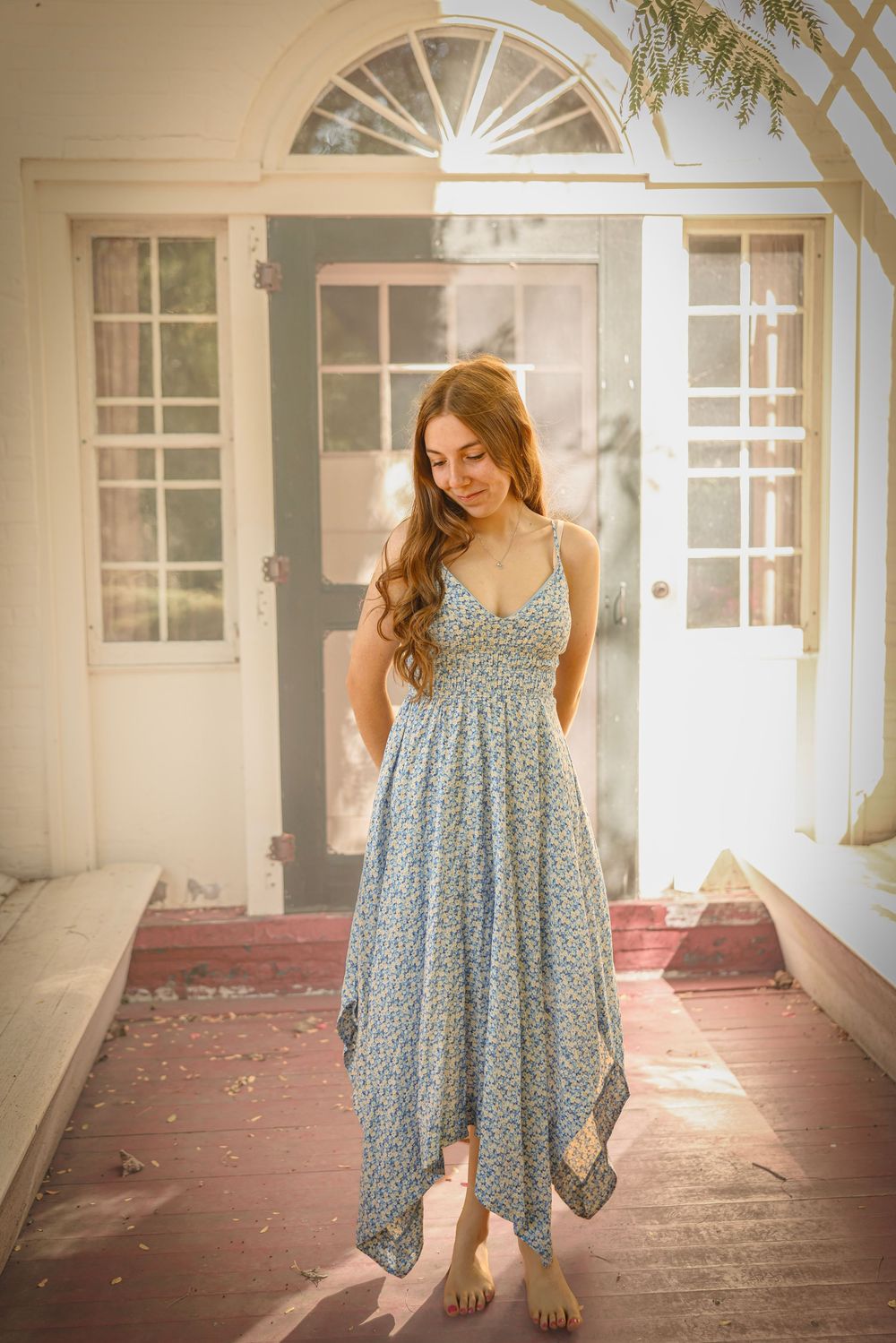 Young woman in a blue floral spaghetti strap maxi dress standing on a sunlit porch.