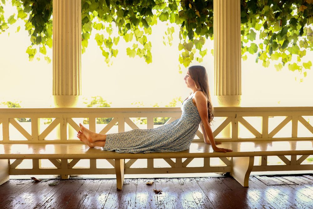 Woman in a floral dress relaxing on a sunlit porch bench surrounded by lush green vines.