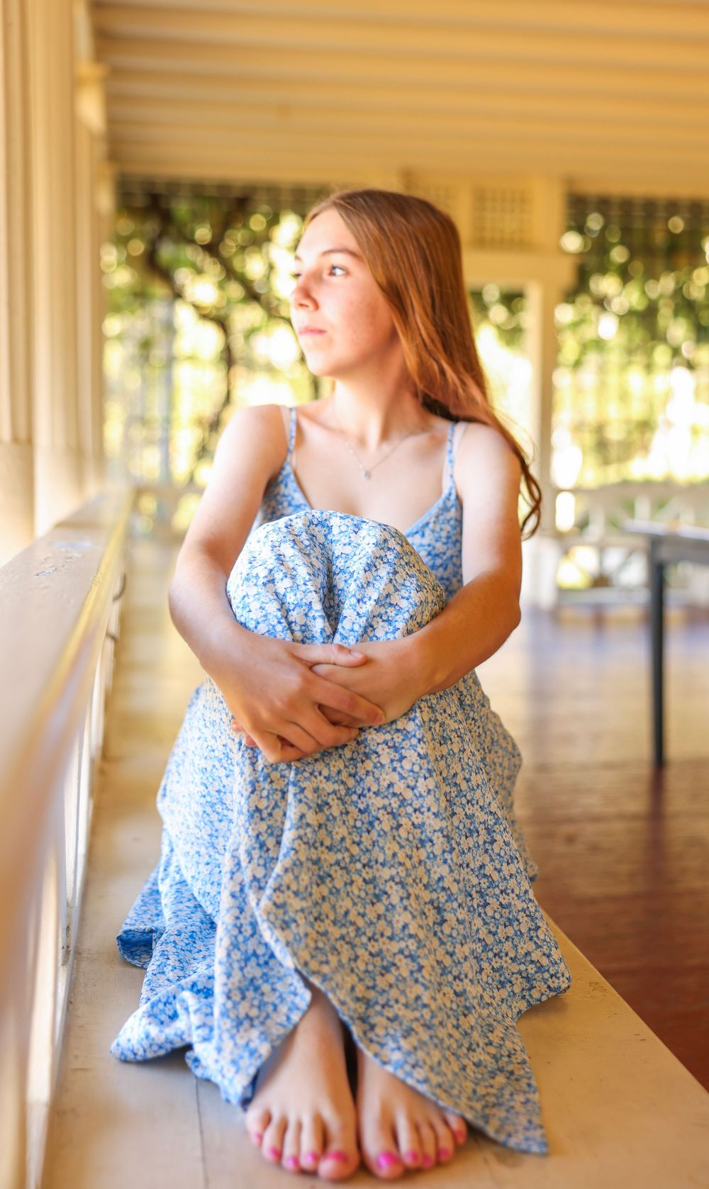 Young woman in a blue floral sundress sitting barefoot on a porch railing, gazing into the distance.