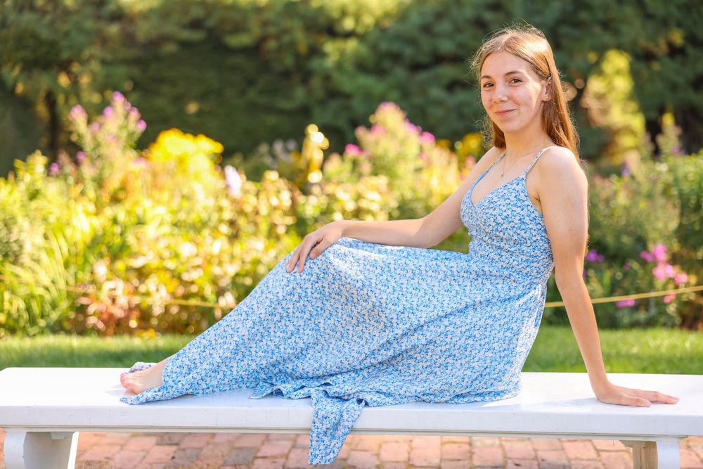 Young woman in a blue floral maxi dress sitting on a white bench in a colorful garden.