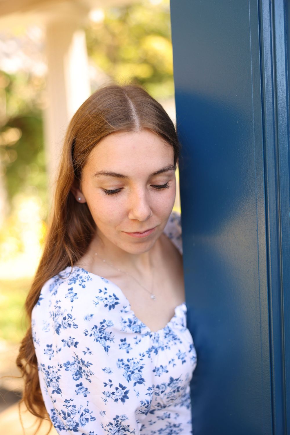 Young woman with long auburn hair in a floral dress leaning against a blue door outdoors.