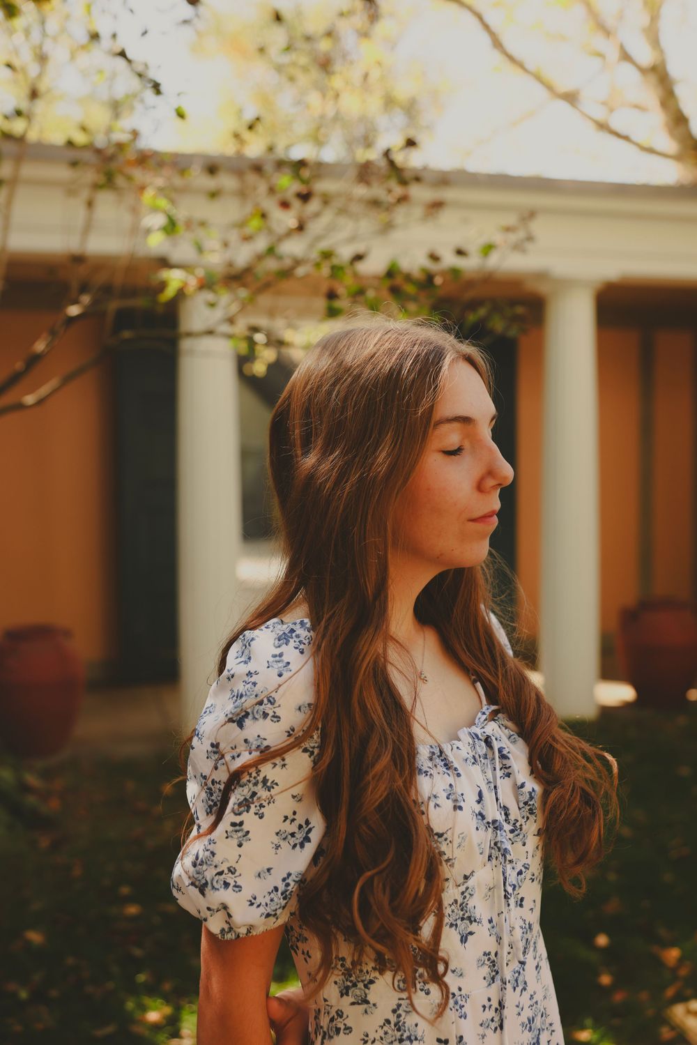 Young woman with long wavy brown hair in a floral dress standing in a sunlit garden, eyes closed.