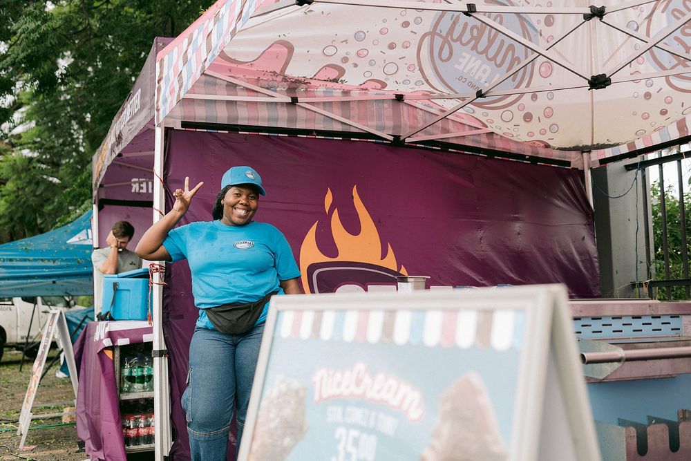 A smiling woman in a blue shirt poses at a colorful food truck stand at an outdoor market event.