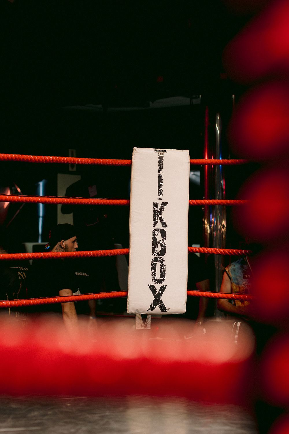 A boxing ring corner post with a white pad labeled TIKBOX, surrounded by red ropes in a dimly lit venue.