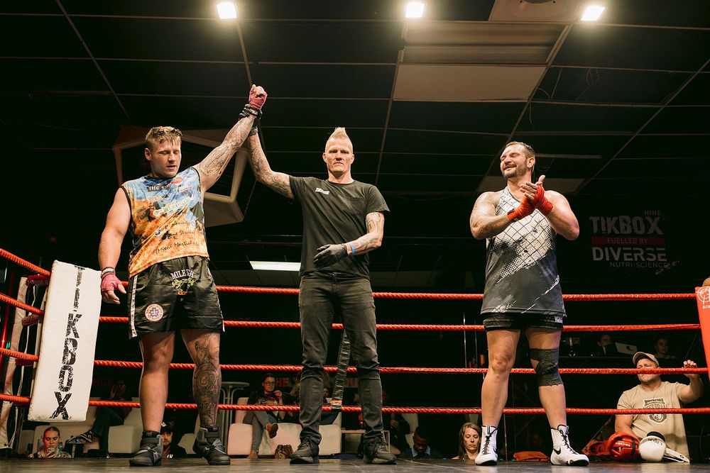 Referee raises winner's hand in kickboxing match between two fighters in a boxing ring at a combat sports event.