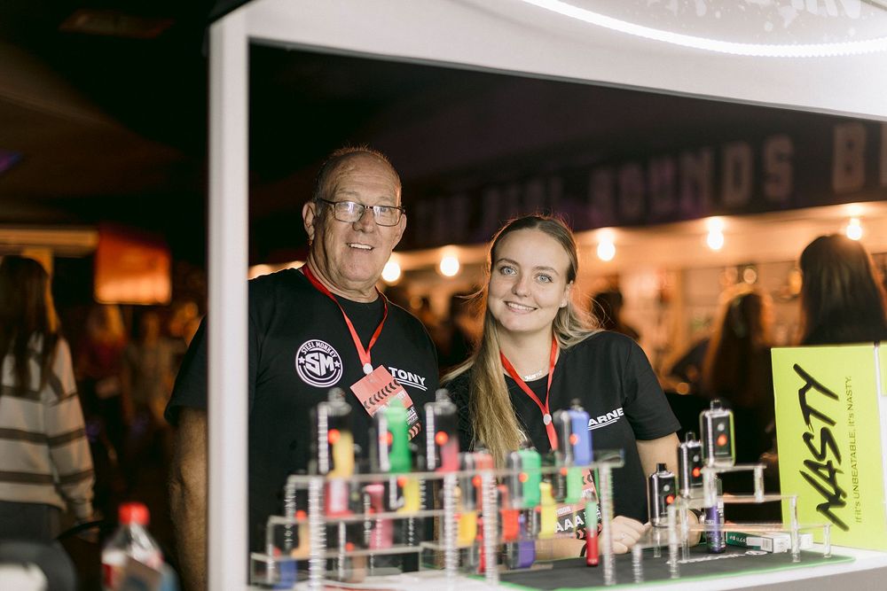 Two smiling staff members pose at a Nasty vape products booth display at an indoor trade event.