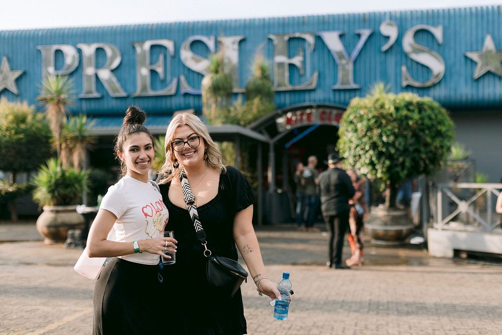 Two women smiling outside Presley's venue, one holding a water bottle, with guests gathered in the background.