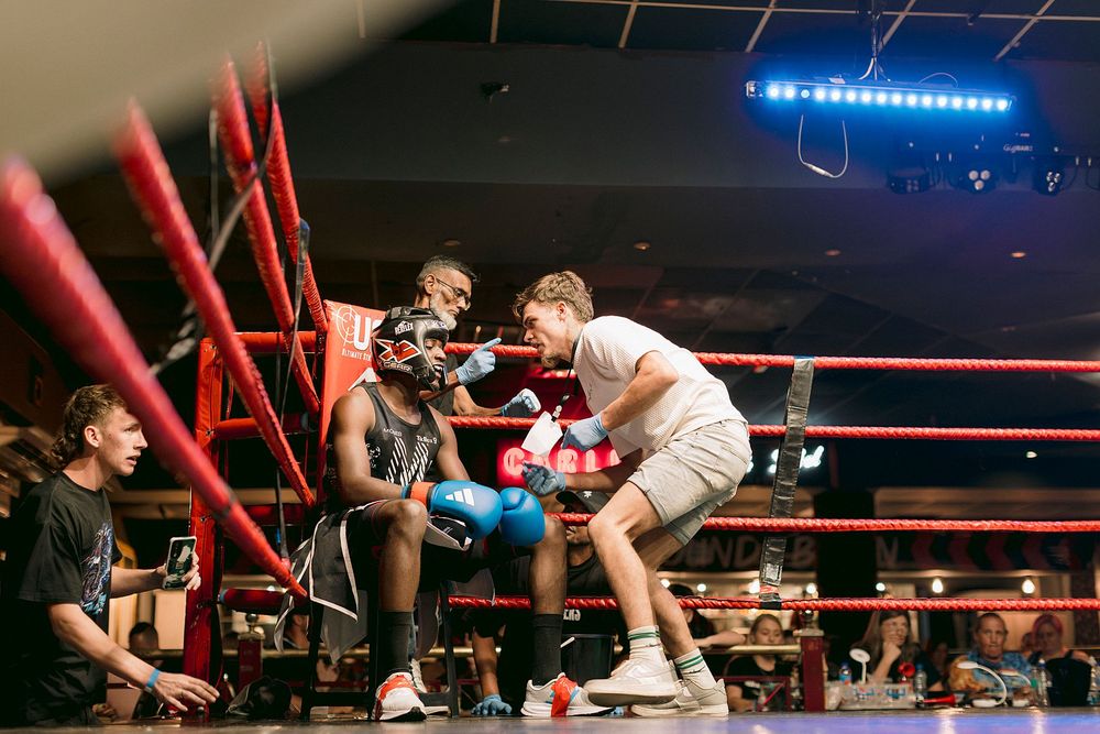 Two boxers sparring in a red boxing ring during an indoor match, with corner men and spectators watching nearby.