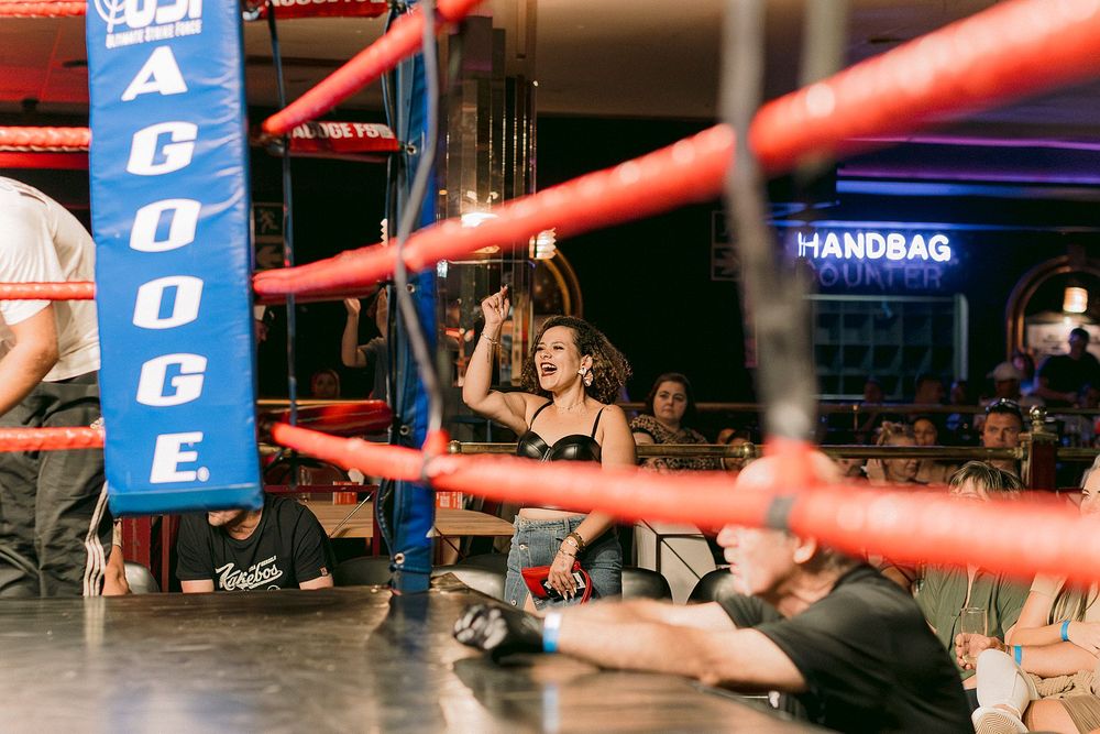 Woman celebrating victory in a boxing ring at a lively venue with red and blue ropes and crowd in background.