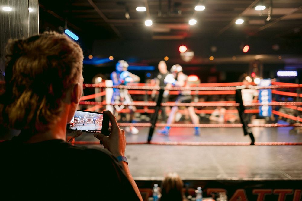 A spectator films a live boxing match on a smartphone ringside at an indoor arena with fighters in the ring.
