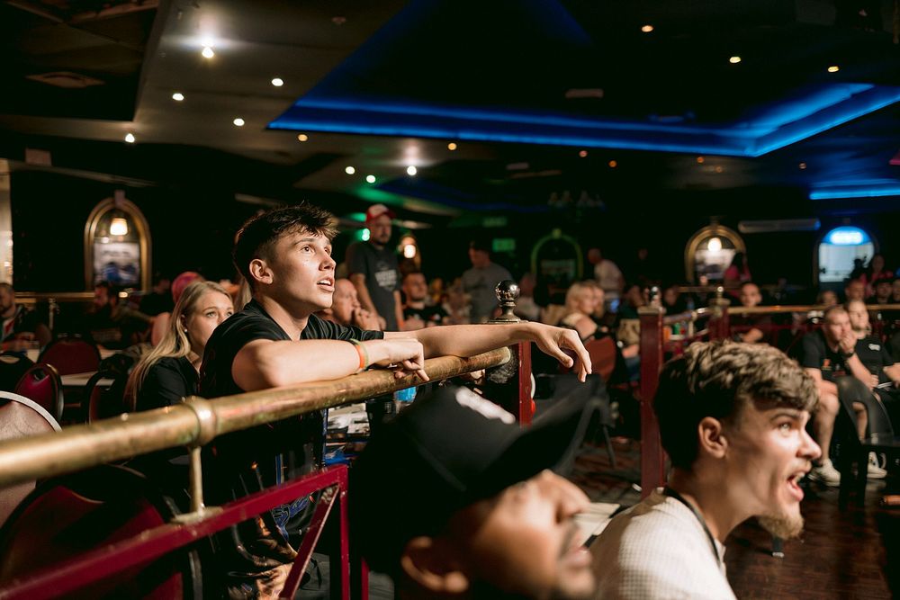 Young crowd leaning on railing at a lively indoor music venue with blue neon lights.