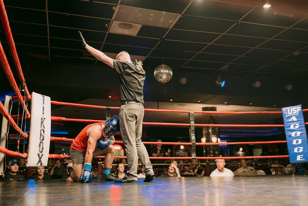 Referee raises hand in boxing ring during USA boxing match, with fighter in red shorts kneeling in corner of roped ring.