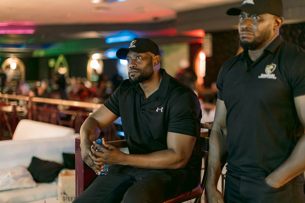 Two men in black polo shirts at an indoor event, one seated with arms crossed, attentively watching something offscreen.