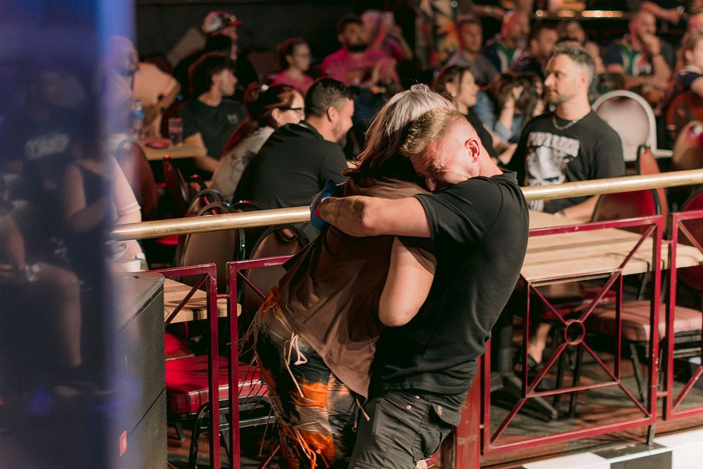 Two people share a warm hug near a railing at a crowded indoor event venue.