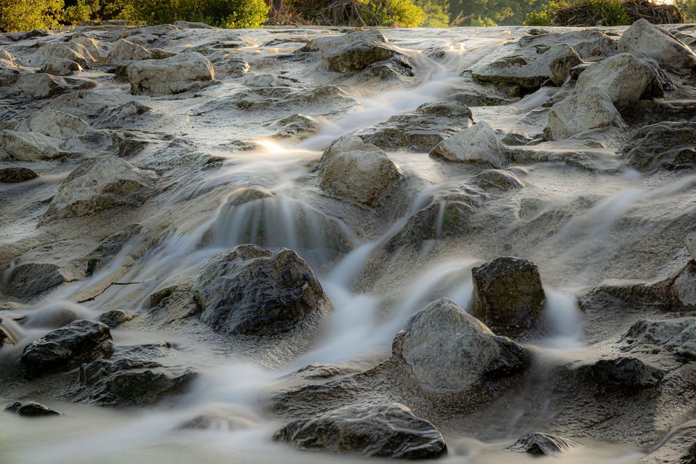 Long exposure photo of silky white water flowing over smooth rocks in a natural rocky riverbed.