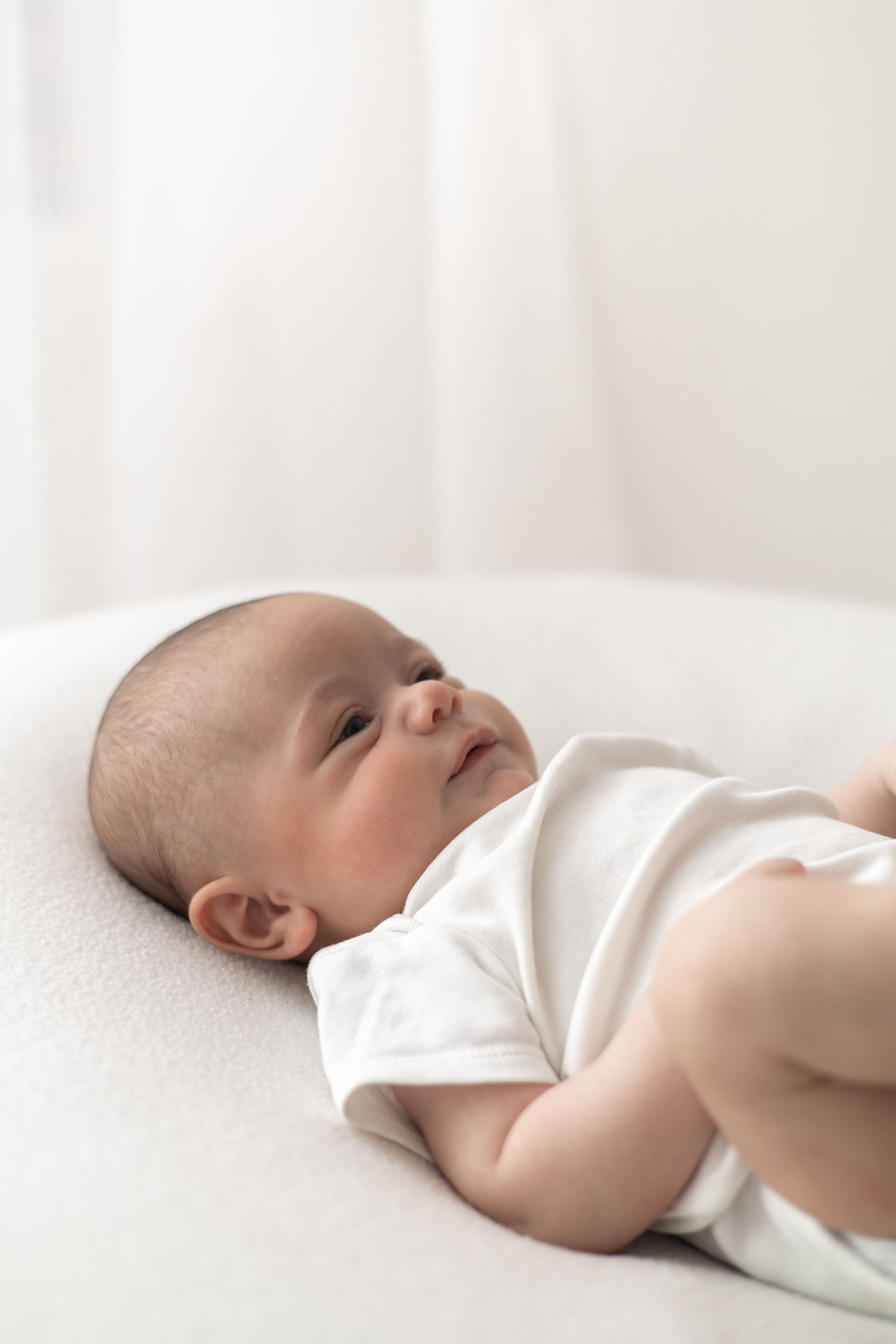 Newborn baby in white onesie lying on soft white surface, looking up natural older newborn baby photography Nottingham