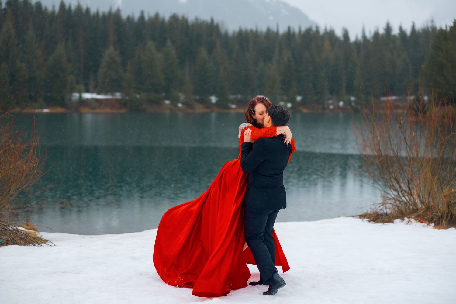 Couple embracing in snow by mountain lake, woman in flowing red gown, winter wedding photo.