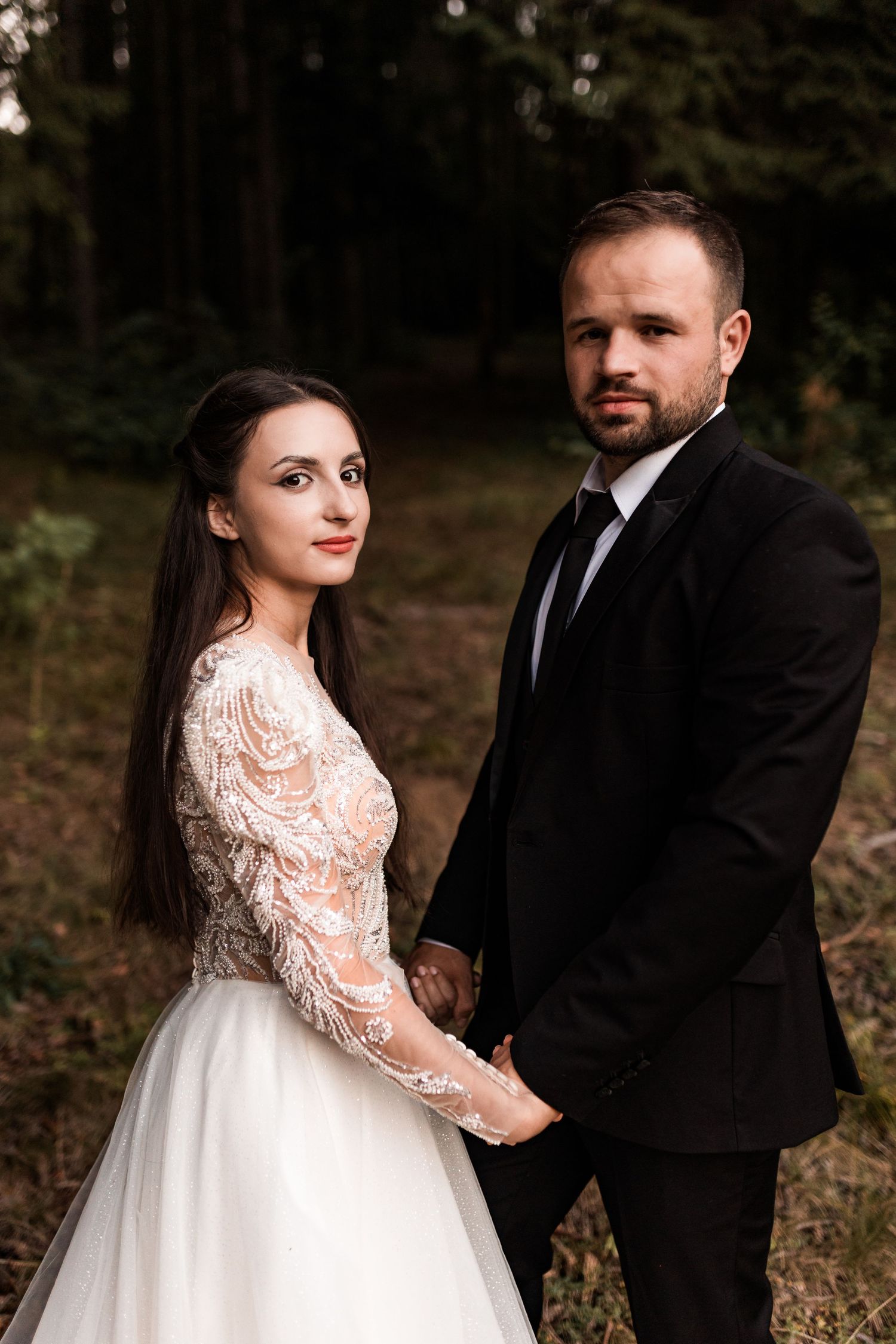 A bride in a lace gown and groom in a black suit hold hands while posing in a dark forest.