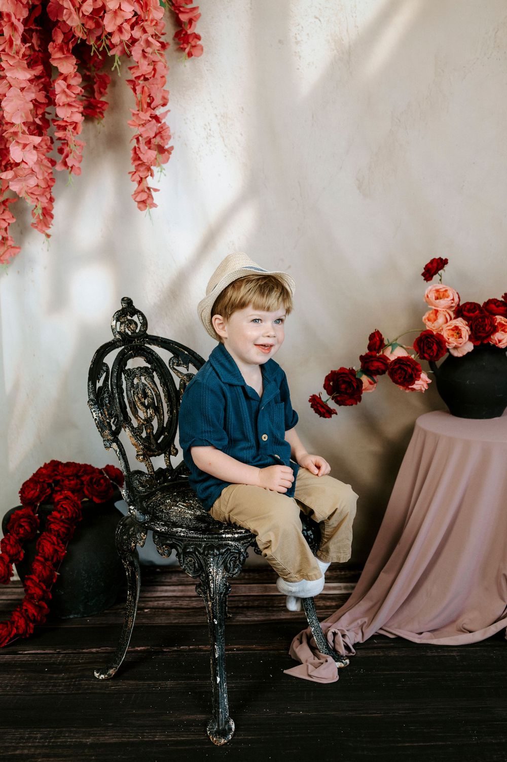 Young boy in blue shirt and hat sits on ornate black chair surrounded by red and pink floral arrangements.