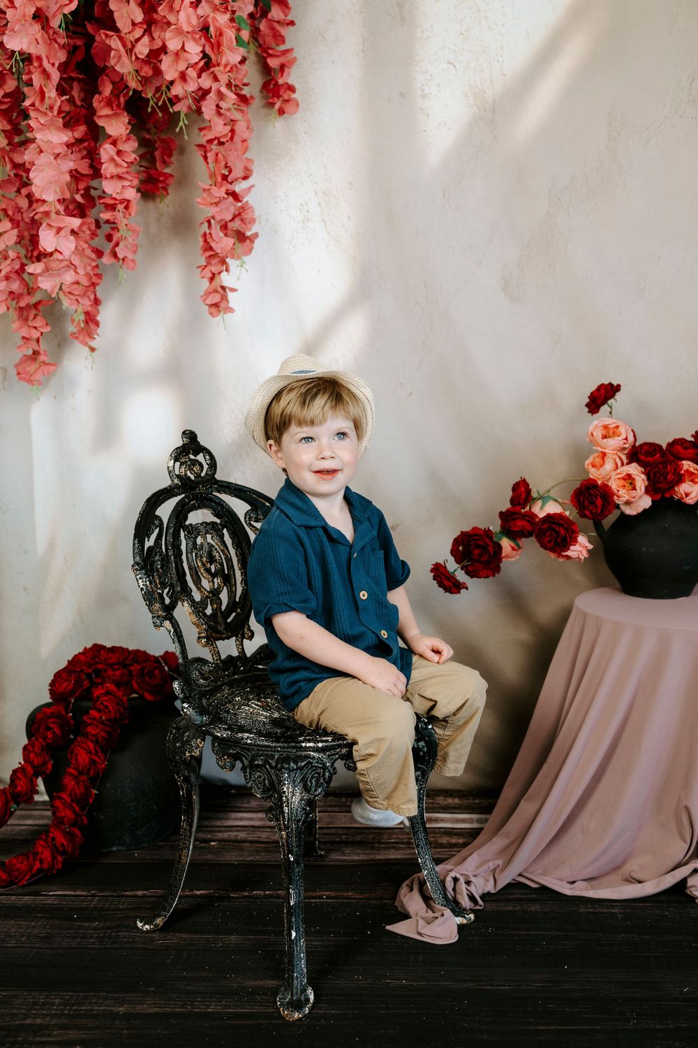 Young boy in navy shirt sits on ornate black chair surrounded by red and pink floral arrangements in studio portrait.