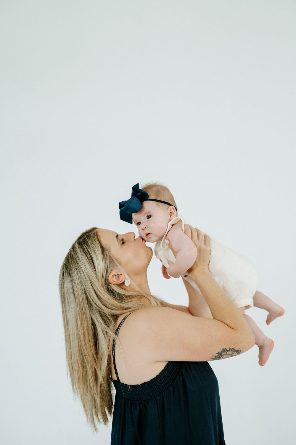 A blonde woman in a black dress kisses her baby girl wearing a navy bow headband against a white background.