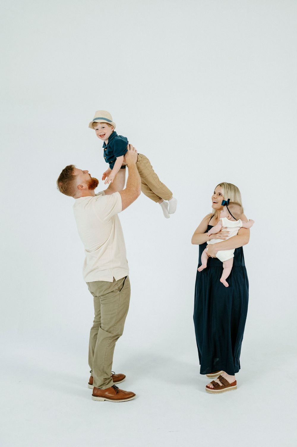 A happy family with a father lifting a toddler boy in the air while mother holds a baby, on white background.