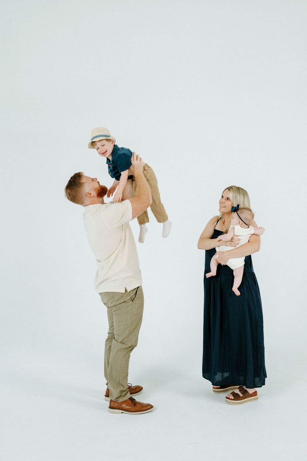 A happy family with a man lifting a toddler in the air while a woman holds an infant against a white background.