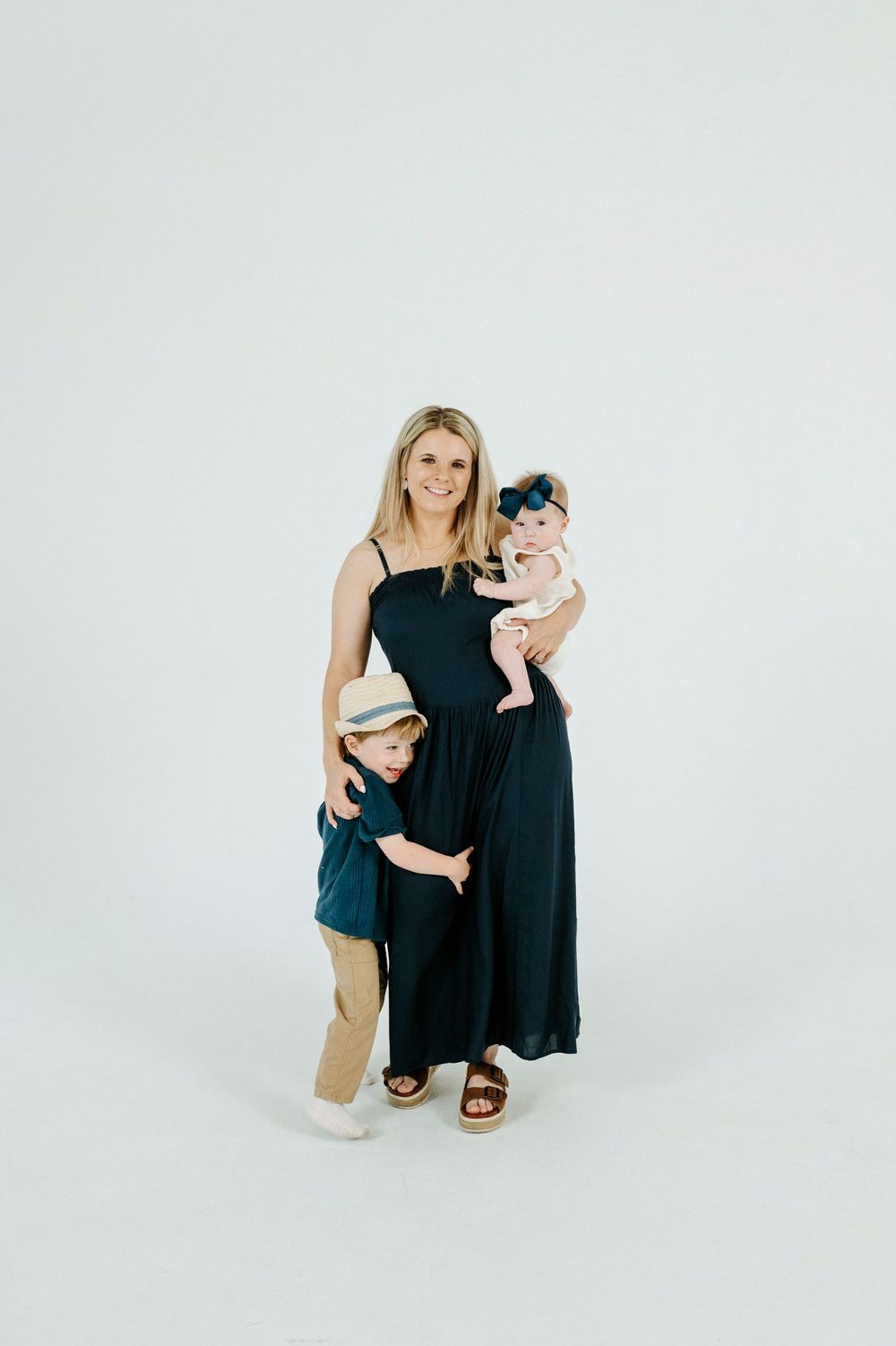 A smiling blonde woman in a navy dress holds a baby girl while a young boy in a hat hugs her side.